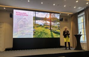 Heli Virkki seisoo näytön edessä, näytöllä teksti "Runkoverkon hyödyt kunnille"