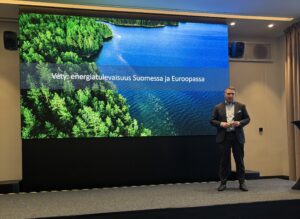 Olli Sipilä seisoo näytön edessä, näytöllä teksti "Vety: energiatulevaisuus Suomessa ja Euroopassa"